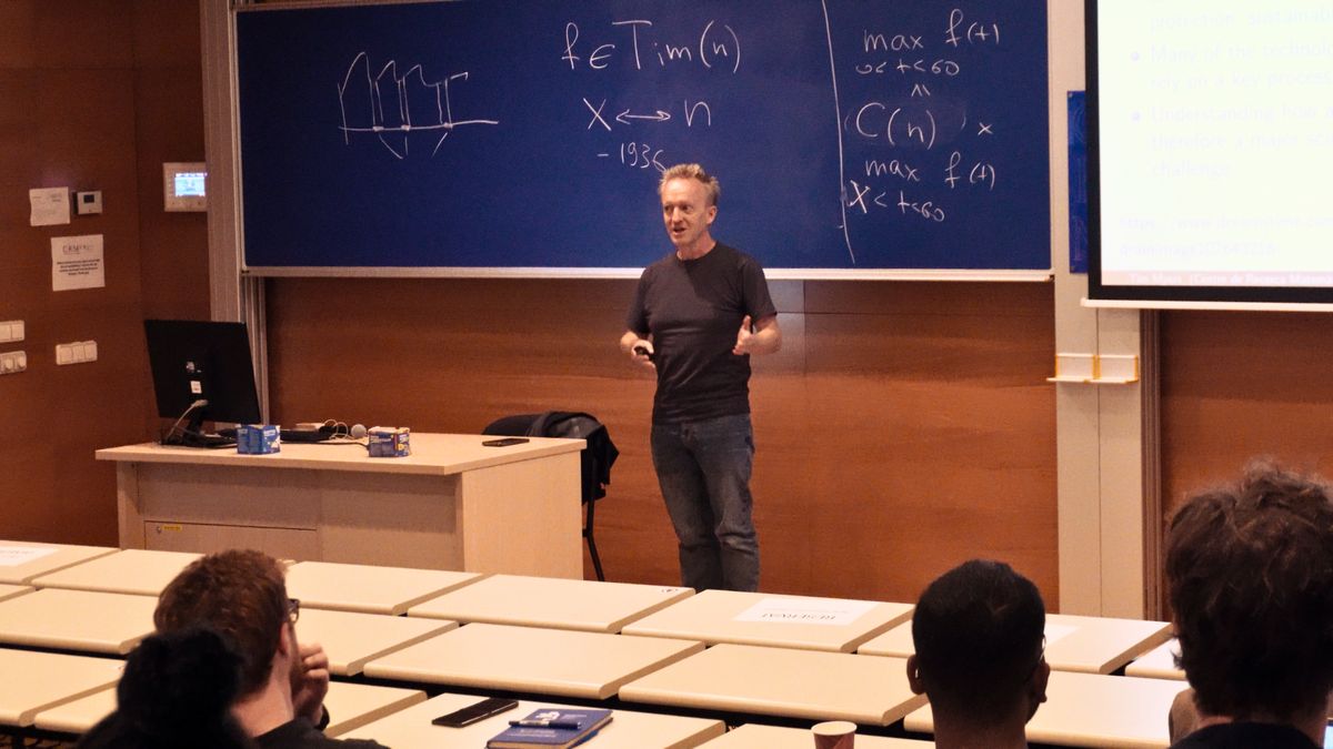Tim Myers speaks to the audience in front of a blackboard with formulas, his slides projected on the right.