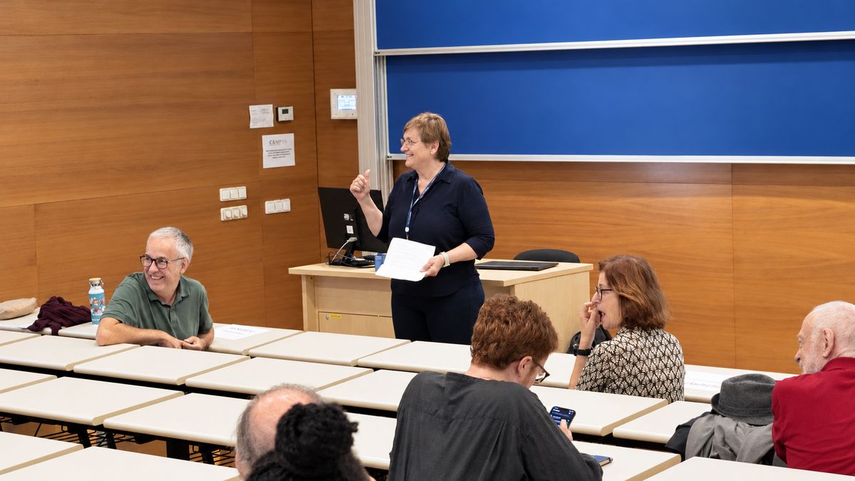 A speaker stands at the front of the lecture hall, holding notes, as people in the front rows listen.