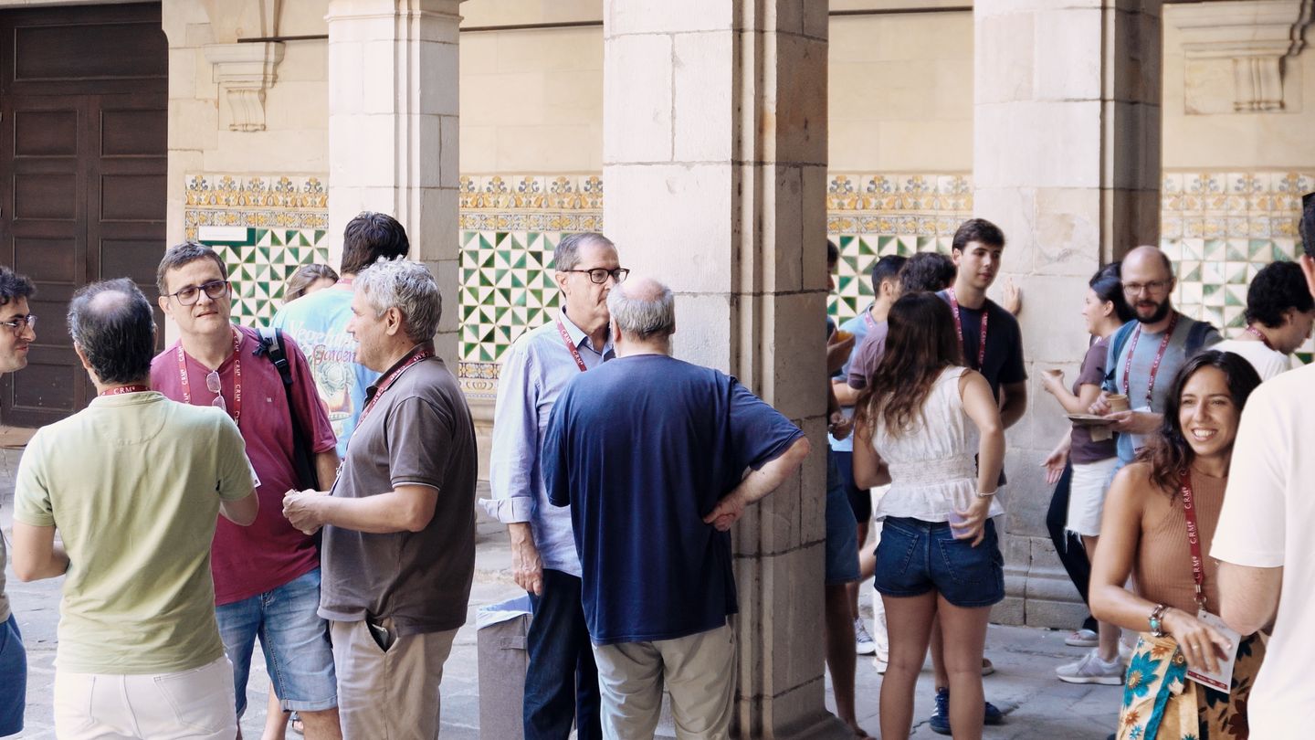 Researchers wearing workshop lanyards talk in small groups under the arches of the cloister, in front of a wall of glazed tiles